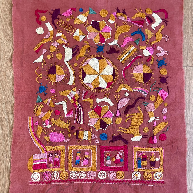 A square pink textile featuring dense, colorful folk-style embroidery with animals, geometric motifs, and a train scene along the bottom, laid flat on a light wood surface.